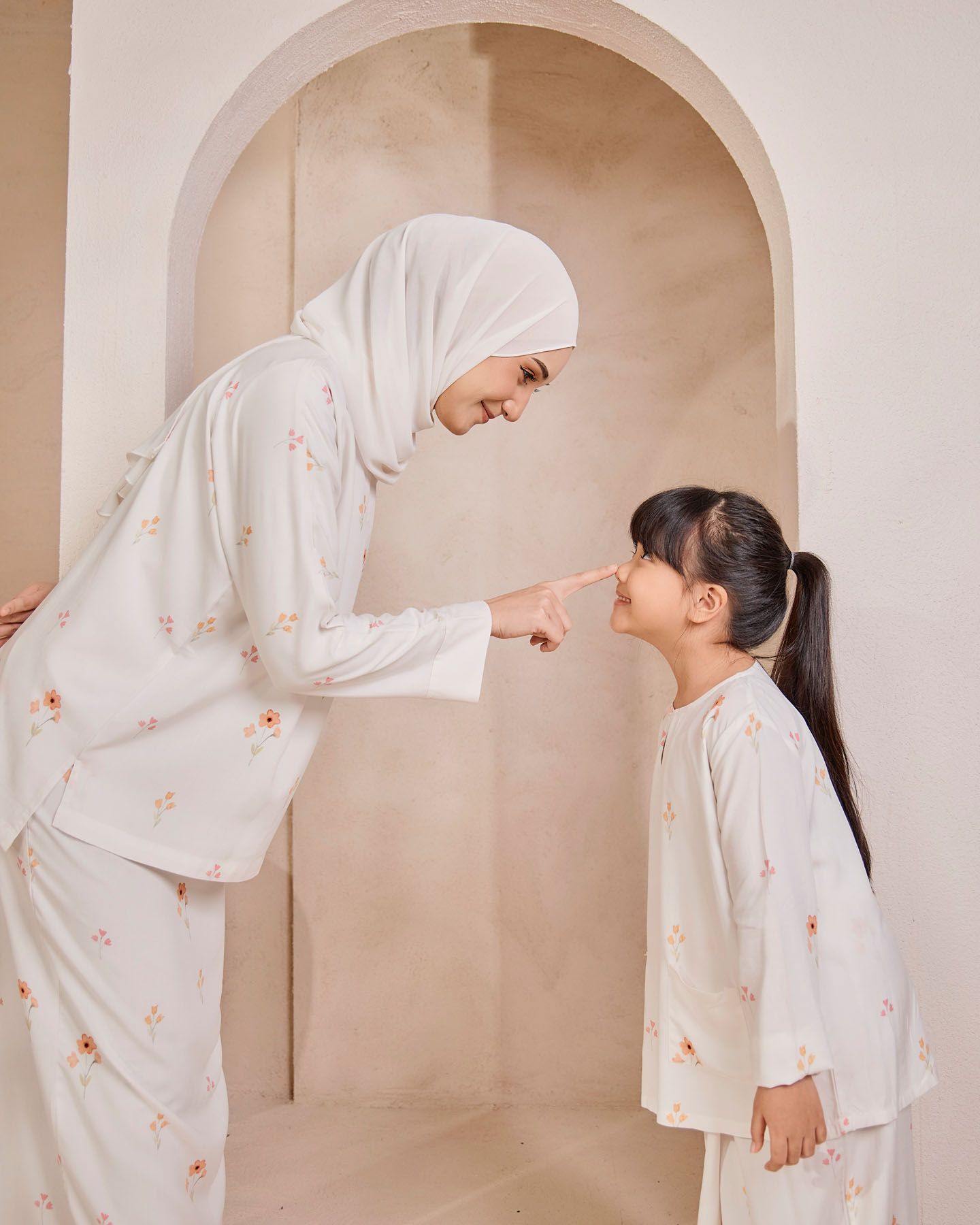 Mother in white floral hijab outfit playfully touching noses with her young daughter in matching attire.
