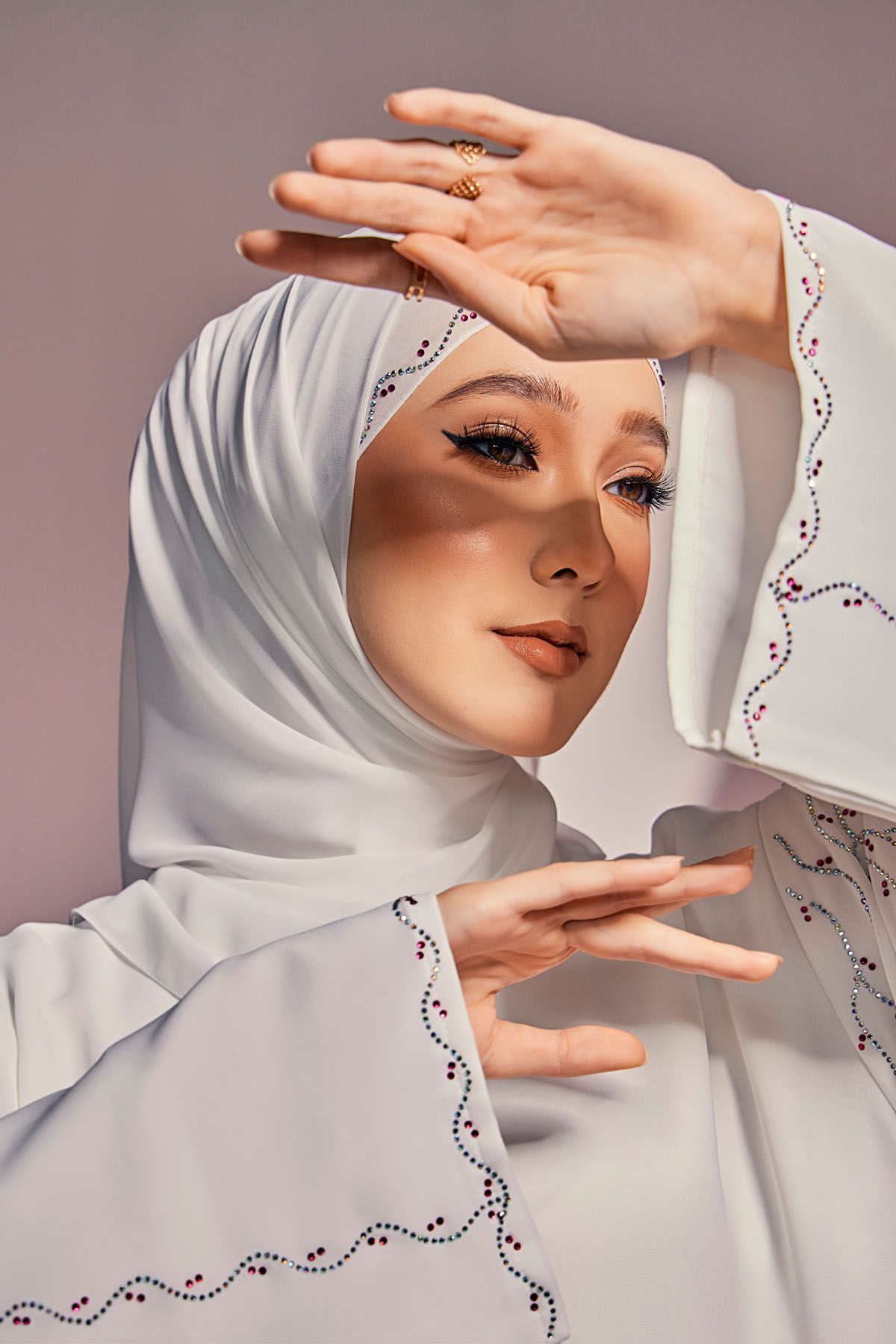 Beautiful woman in white embroidered hijab and abaya framing her face with both hands gracefully.