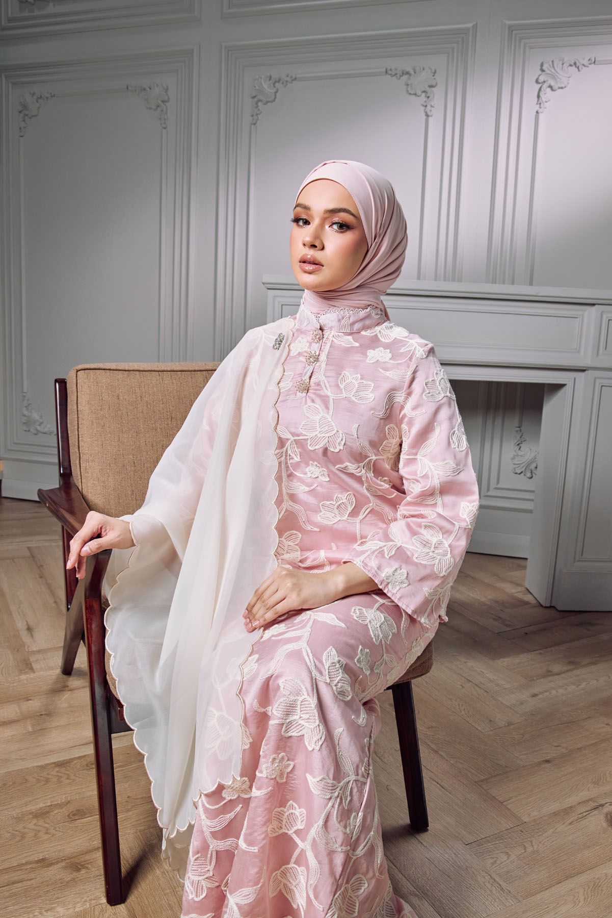 Elegant Muslim woman in pink floral kebaya and hijab seated on chair in white room.