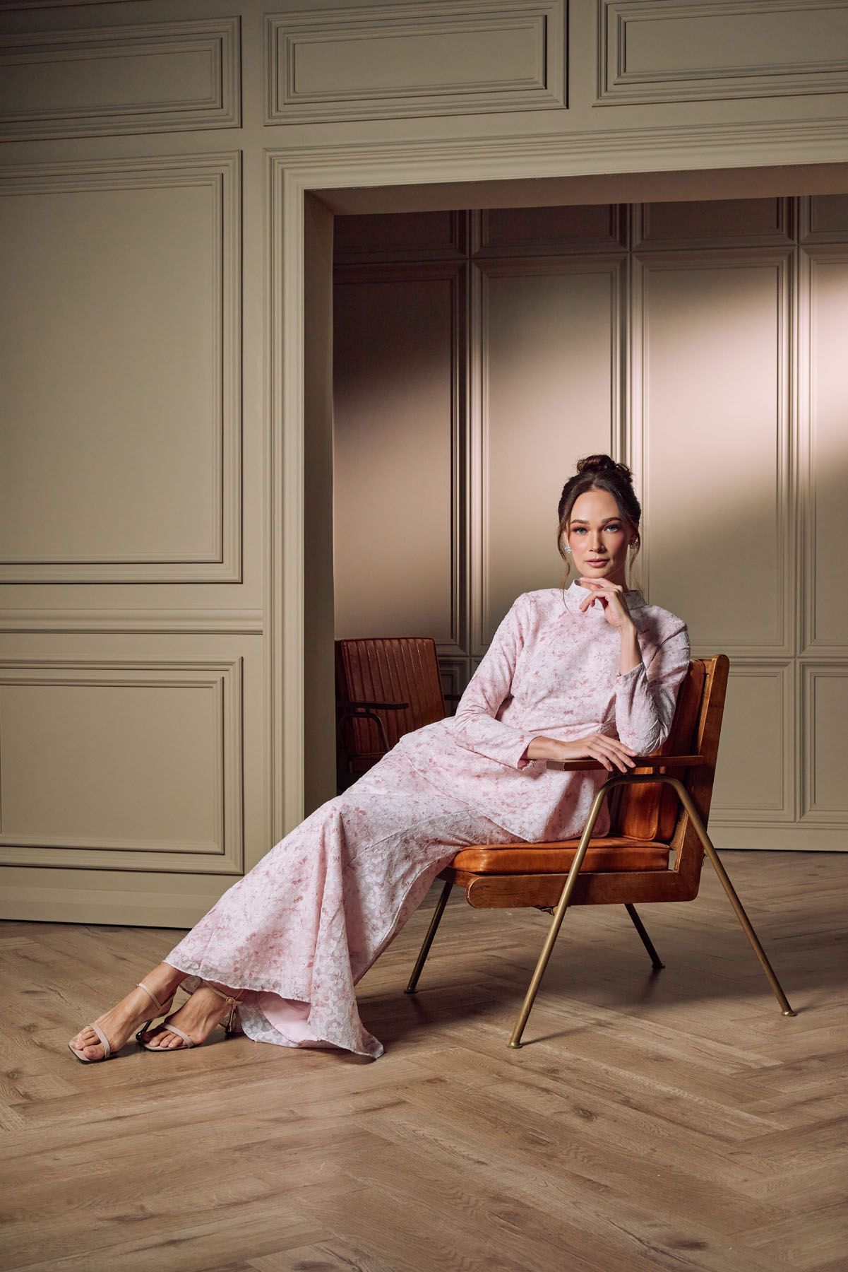 Woman in pink floral modest dress sits on leather chair in elegant room.
