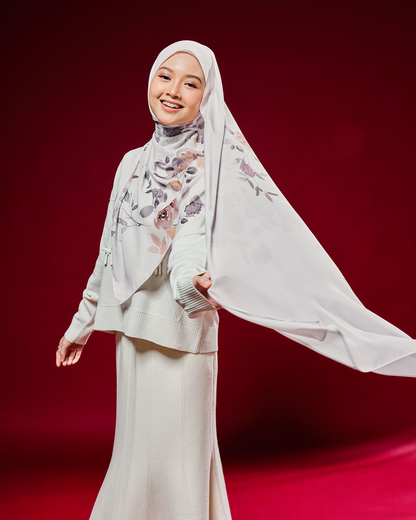 Woman in white floral hijab and cream baju kurung twirls elegantly on red background.