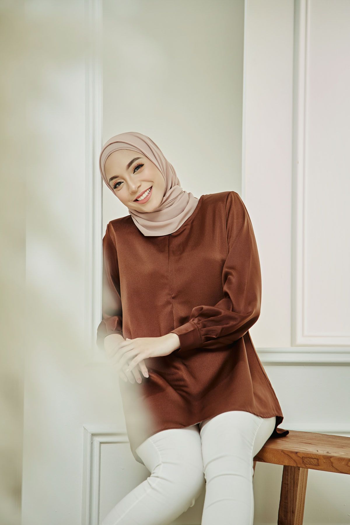 Smiling woman in brown satin tunic and beige hijab sitting on wooden bench in bright white room.