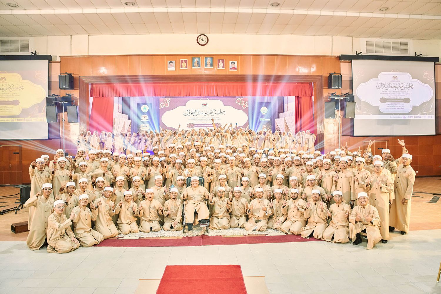 Large group photo of students and officials in matching beige uniforms at a Malaysian Quran memorization ceremony.