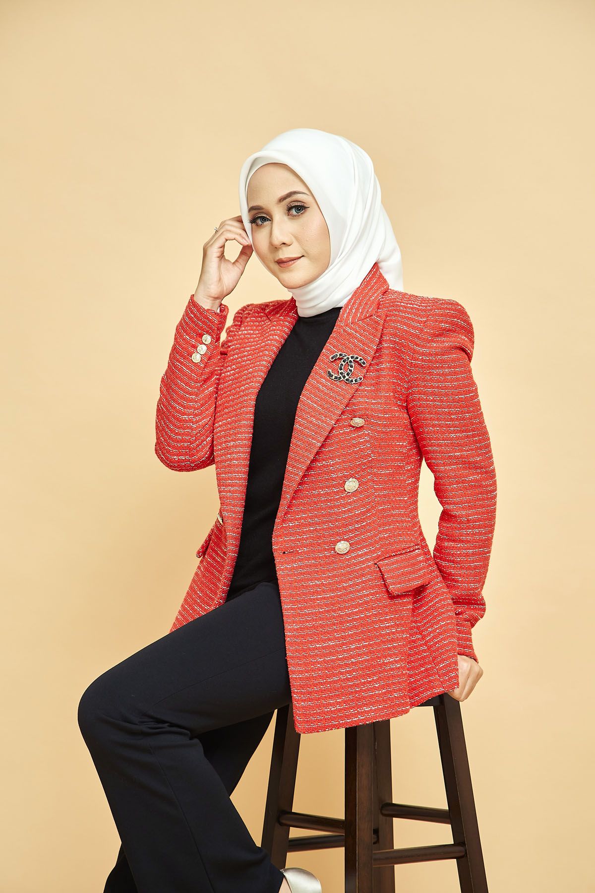 Elegant Muslim woman in white hijab and red tweed blazer seated on stool against beige backdrop.
