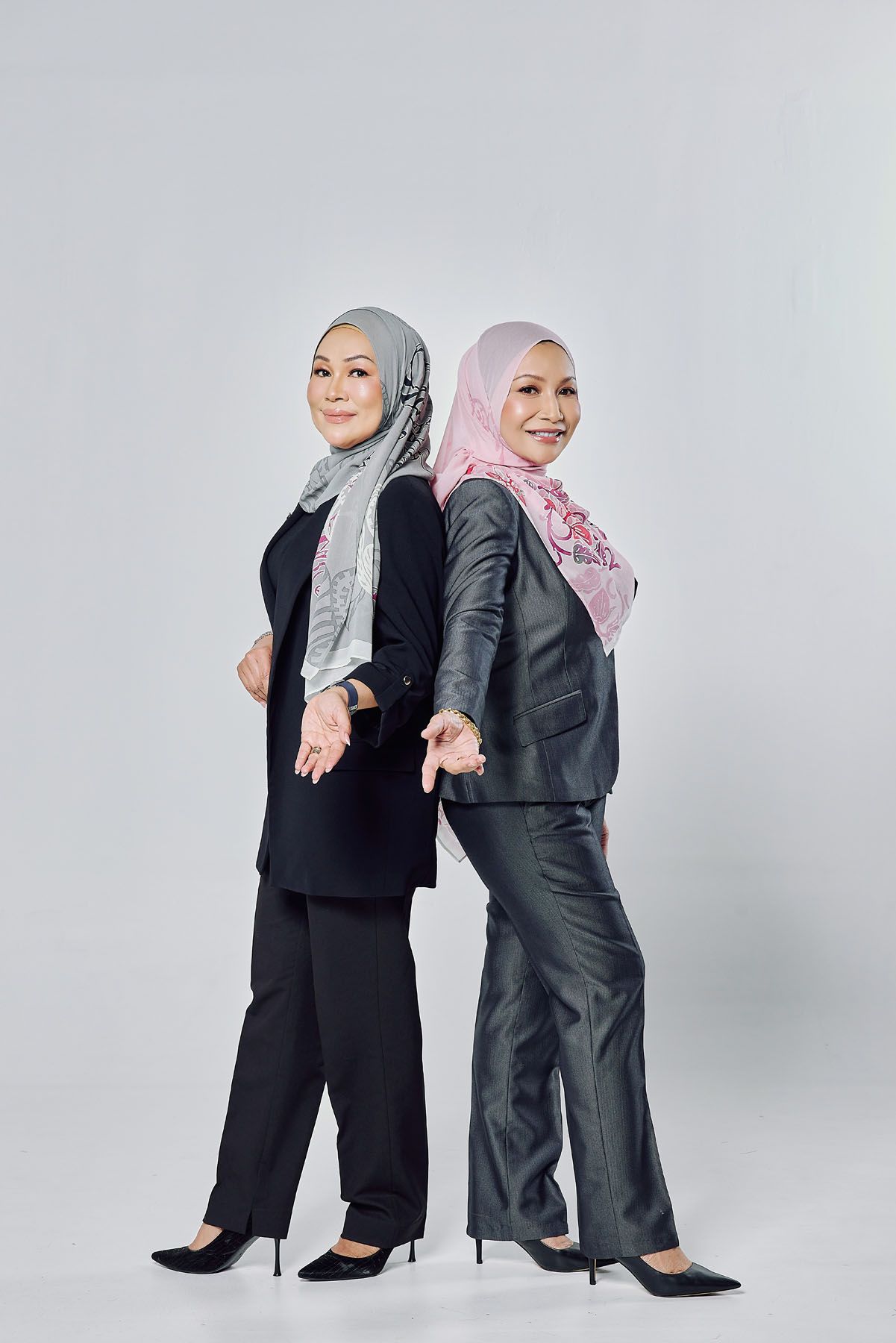 Two professional Muslim women in hijabs and dark business suits posing back-to-back in a studio.