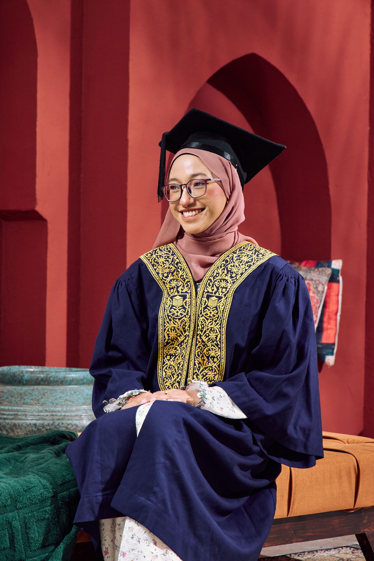 Smiling Muslim graduate in navy embroidered gown and pink hijab poses before red Moroccan arched walls.