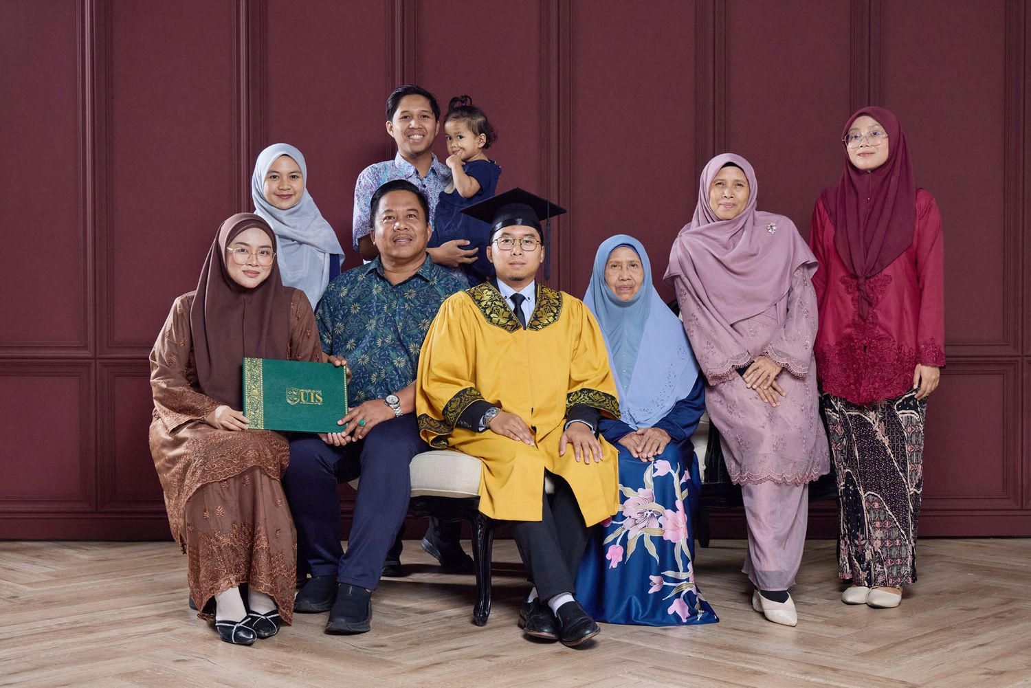 Graduate in yellow and blue academic robes sits with family group of eight people before a dark red wall.