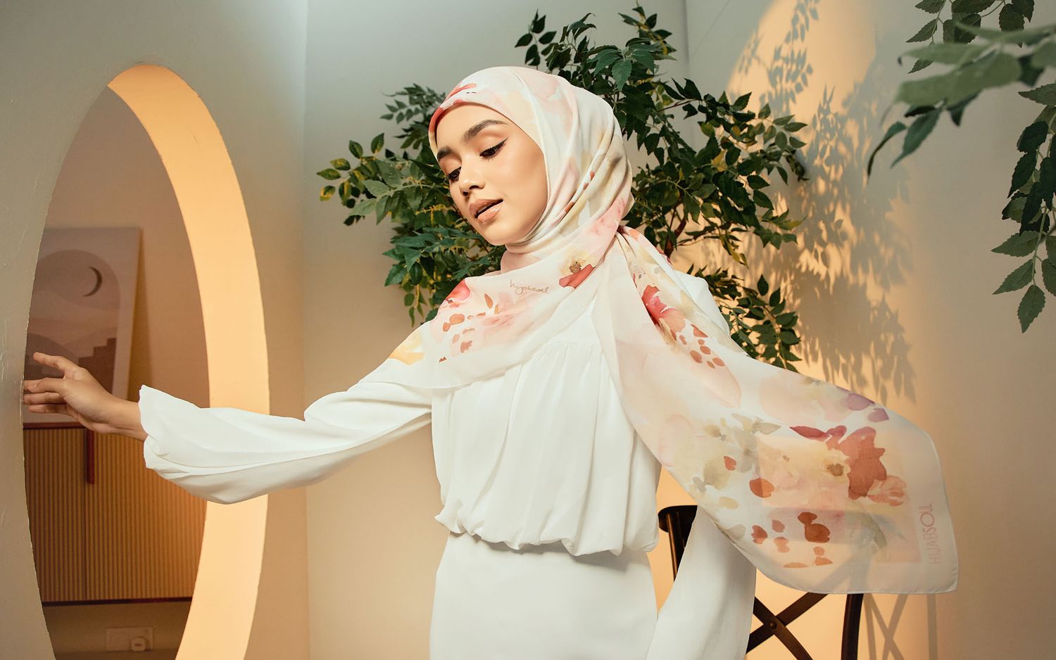 Woman in a white dress with a floral chiffon hijab posing gracefully in an archway setting.