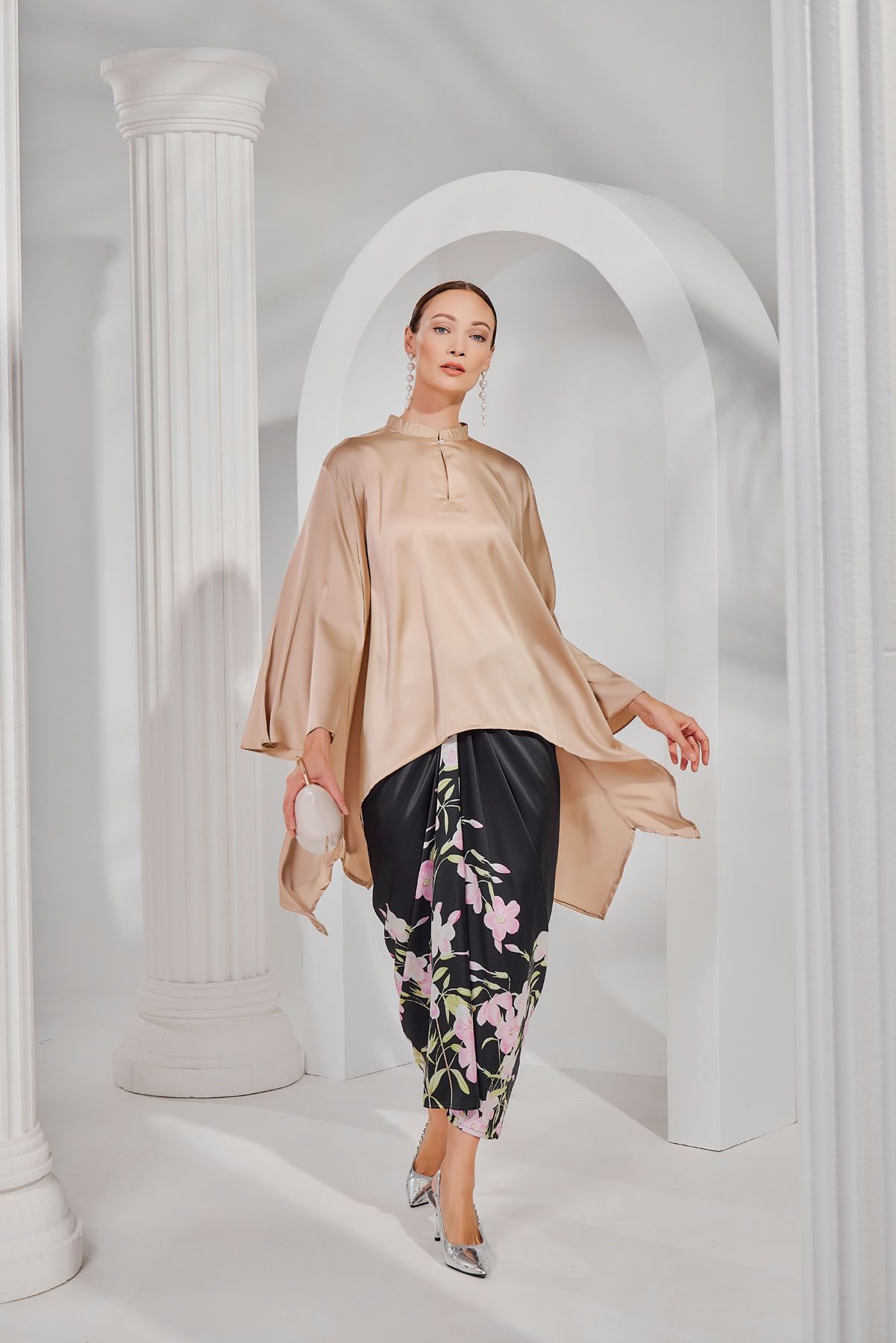 A woman in a beige satin kaftan top and floral black pants poses near white arched columns.
