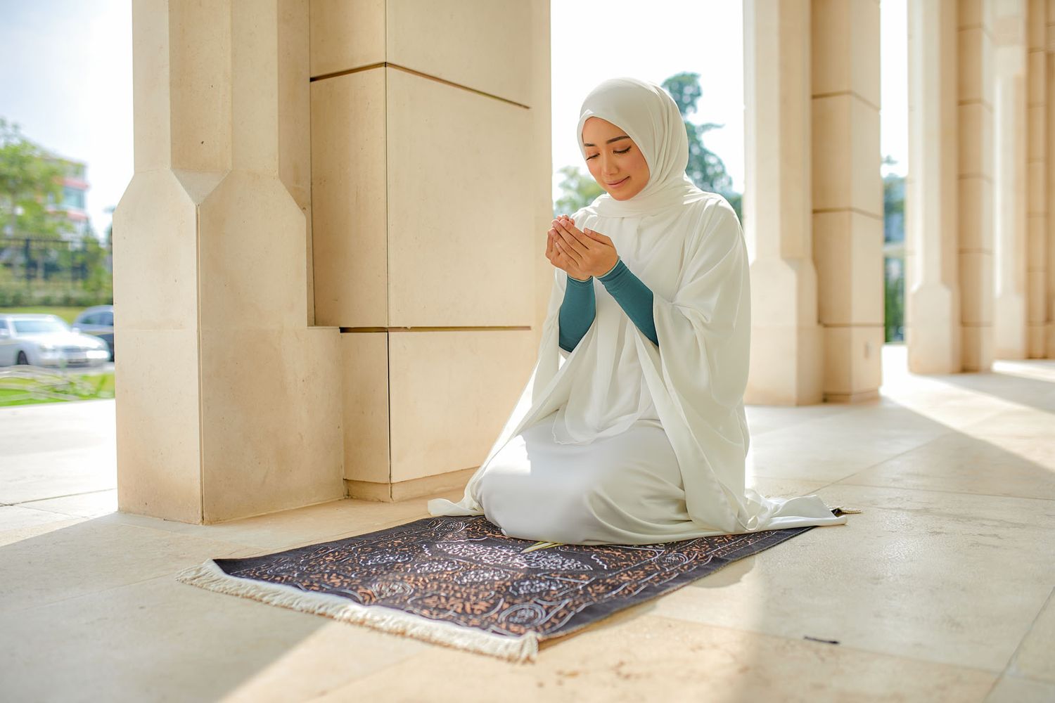 Muslim woman in white hijab praying on decorative prayer mat at sunlit mosque colonnade.