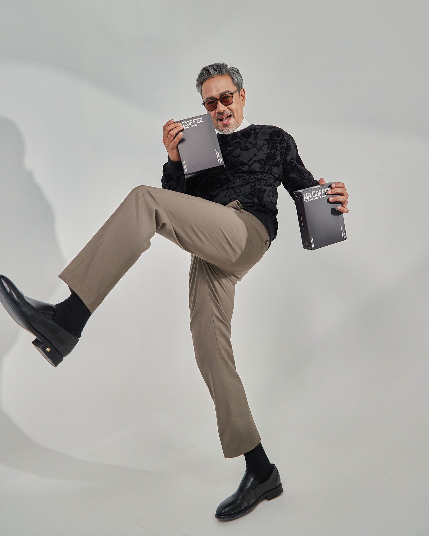 Energetic older man in black sweater and khaki pants kicks leg up while holding Mr. Coffee books.