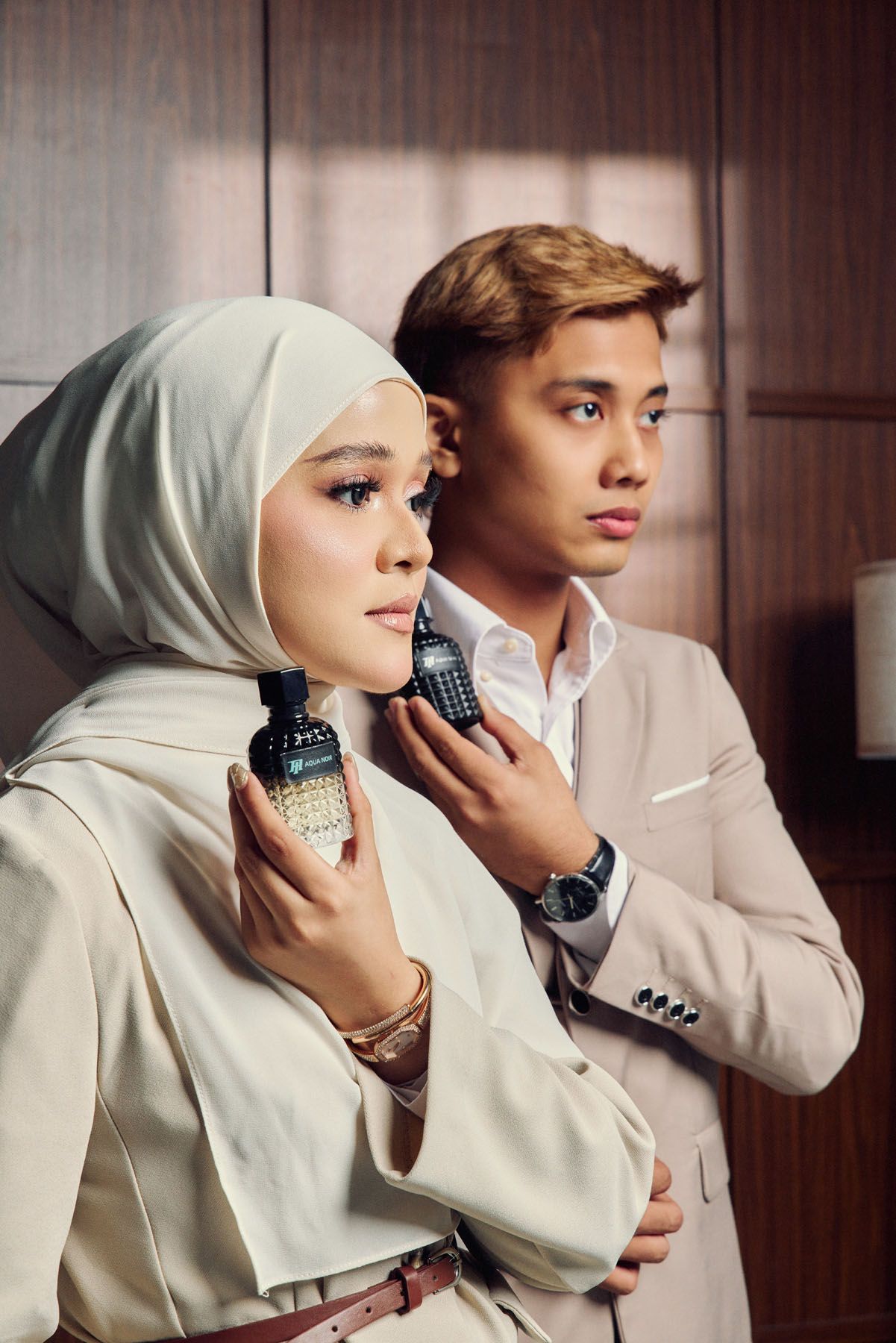 Muslim couple in beige outfits holding dark perfume bottles in elegant wooden interior.