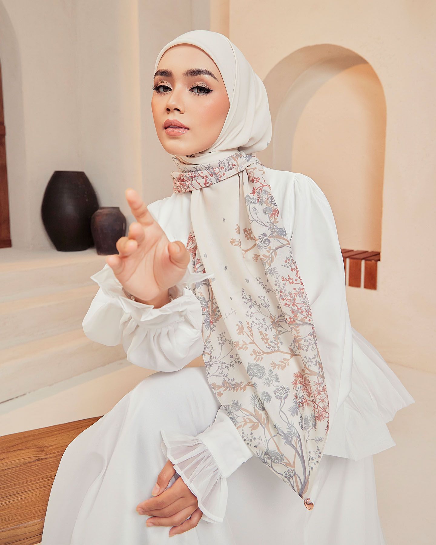 A woman in a white hijab and white dress wears a delicate floral printed scarf in a softly lit arched interior.
