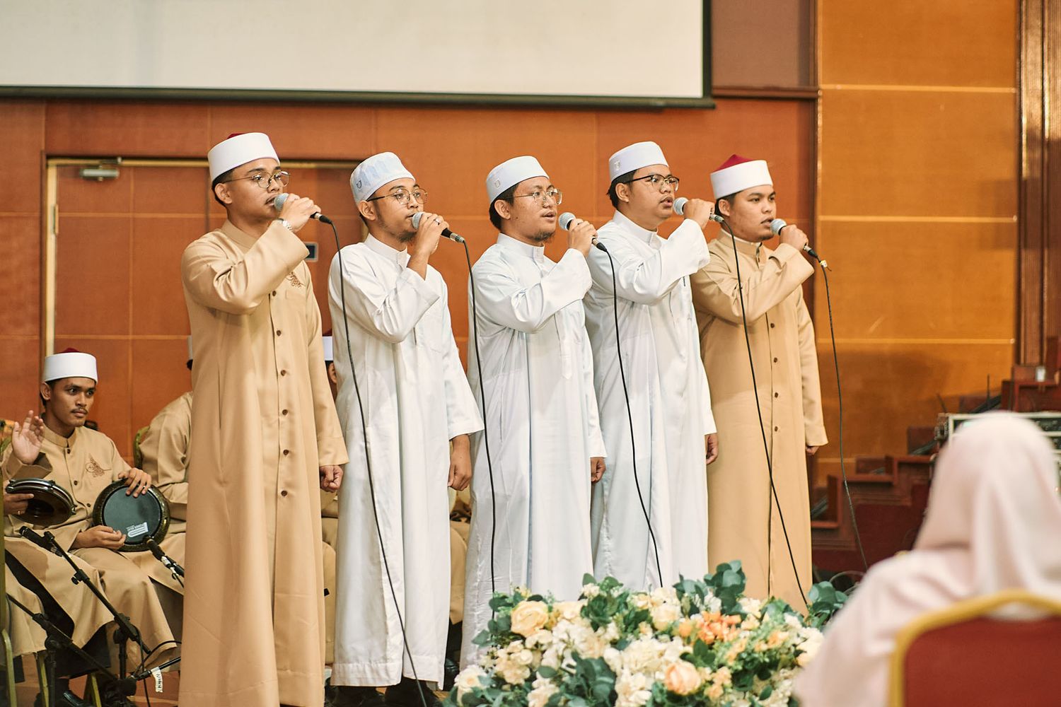 Five Muslim men in white robes and caps performing nasheed vocal performance at an Islamic event.