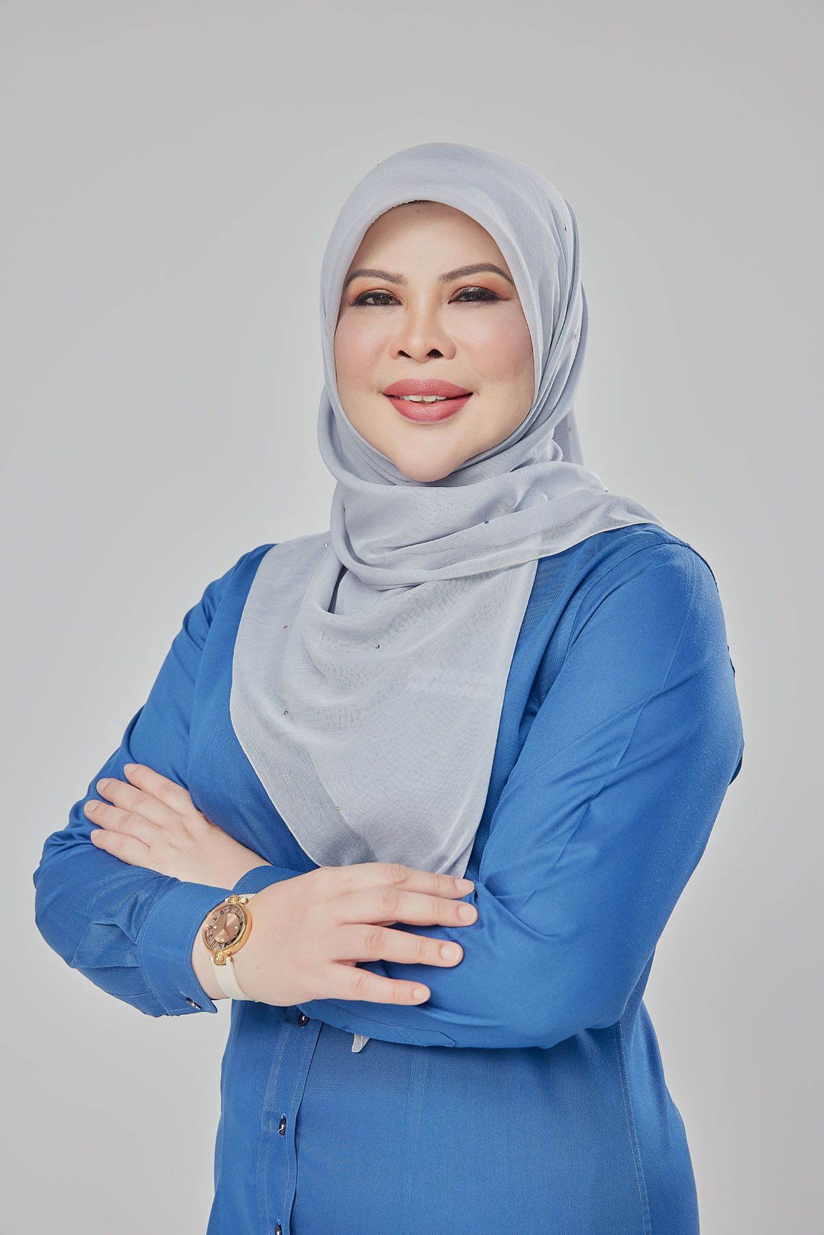 Professional Muslim woman in blue blouse and grey hijab posing with arms crossed confidently.
