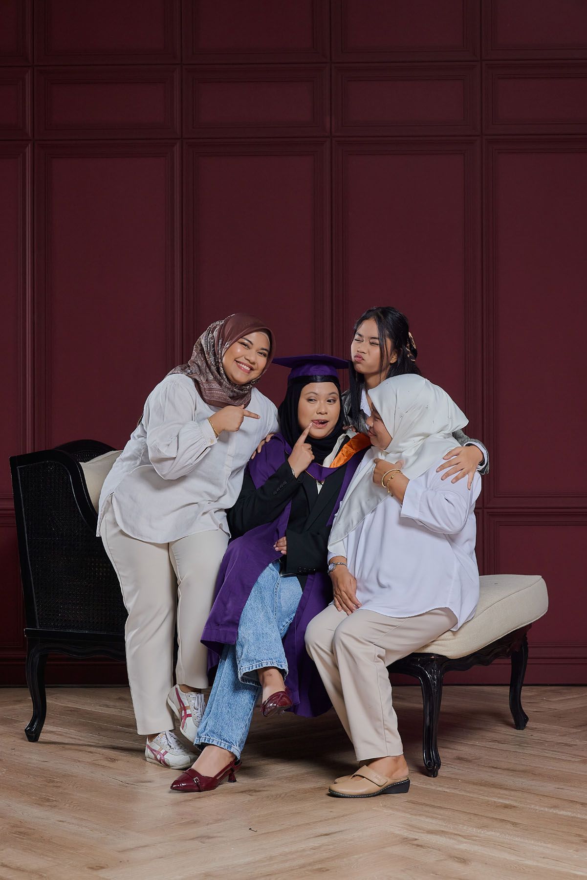 Three women posing joyfully with graduate in purple cap and gown on elegant bench.