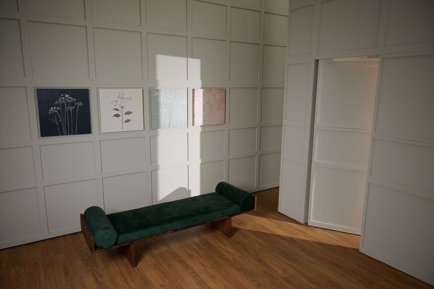 A minimalist gallery room with a green velvet bench, wood floor, white paneled walls, and two botanical art prints displayed.