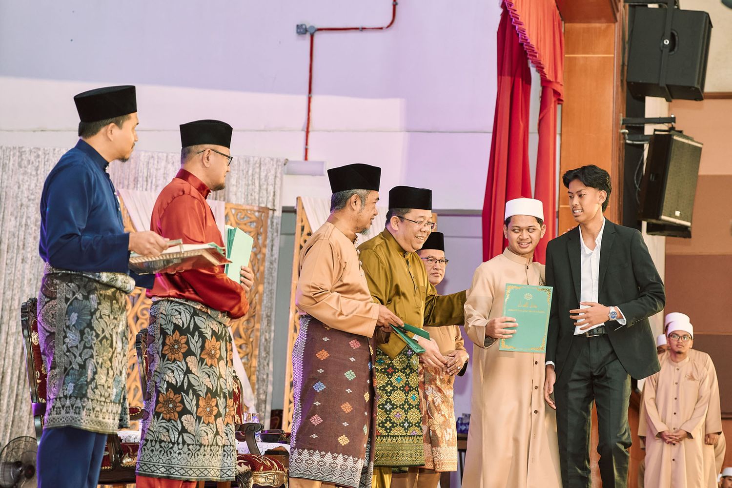 Group of Malaysian officials in traditional attire presenting certificates to recipients at a formal Islamic event.
