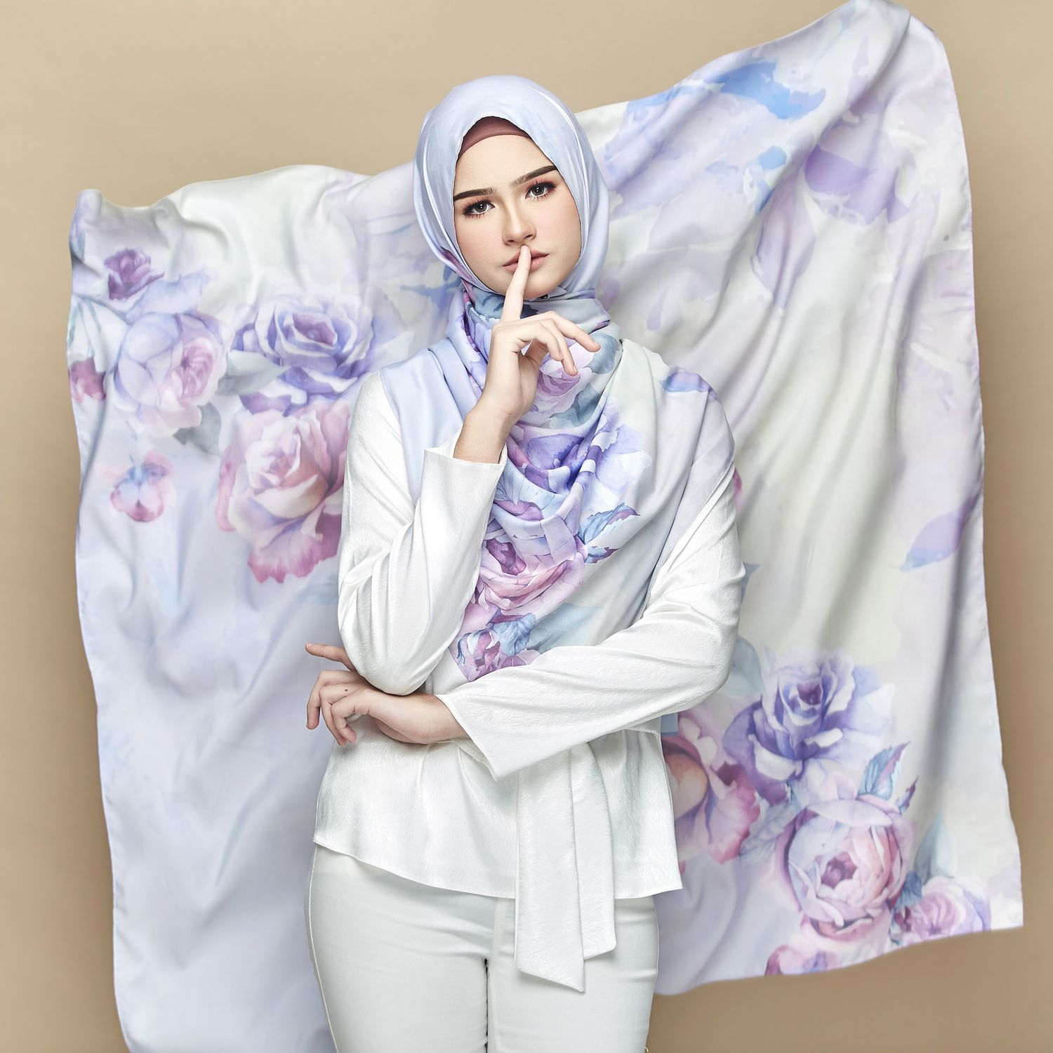Woman in white hijab and floral printed scarf posing with finger to lips against neutral background.