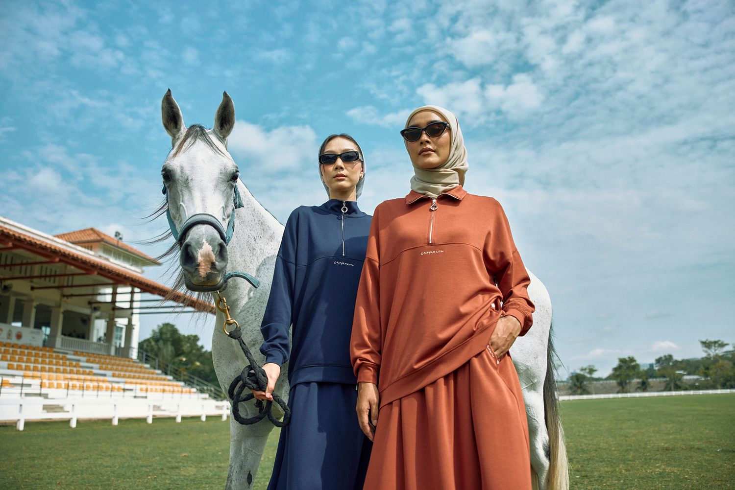 Two women in navy and rust modest outfits with white horse at equestrian stadium.