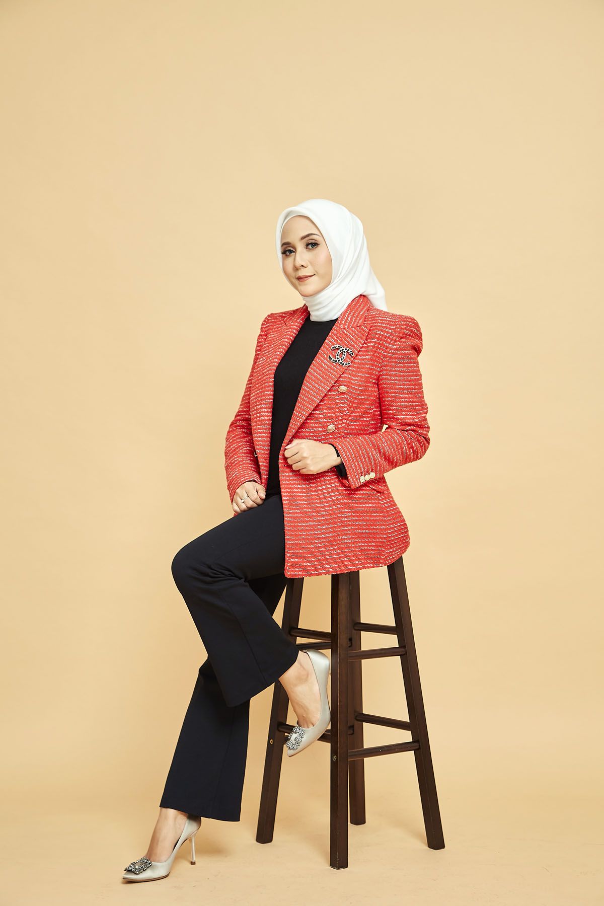 Muslim woman in white hijab and red tweed blazer sitting on wooden stool on beige background.