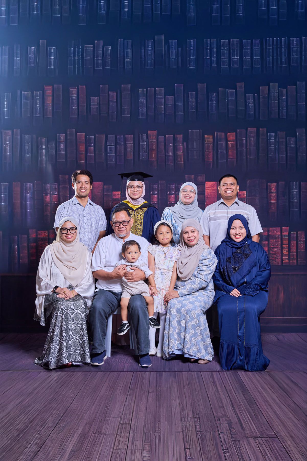 Large family group portrait with graduate in blue cap and gown centered among nine relatives before bookshelf backdrop.