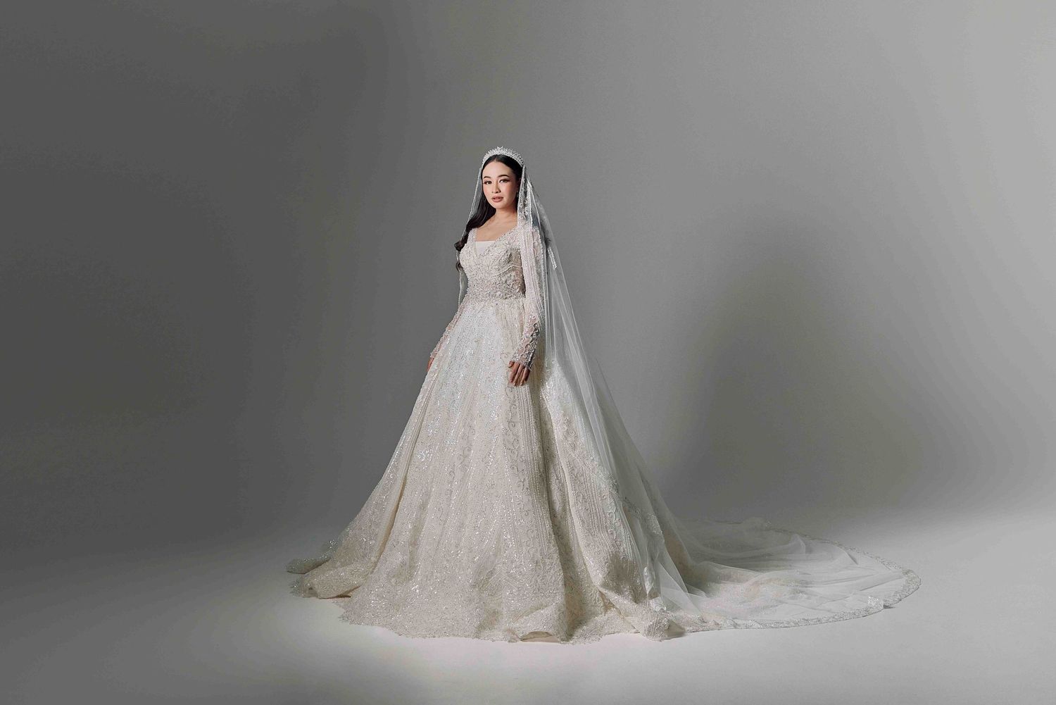 Bride wearing an elegant long-sleeve white lace ball gown with a cathedral veil on grey background.