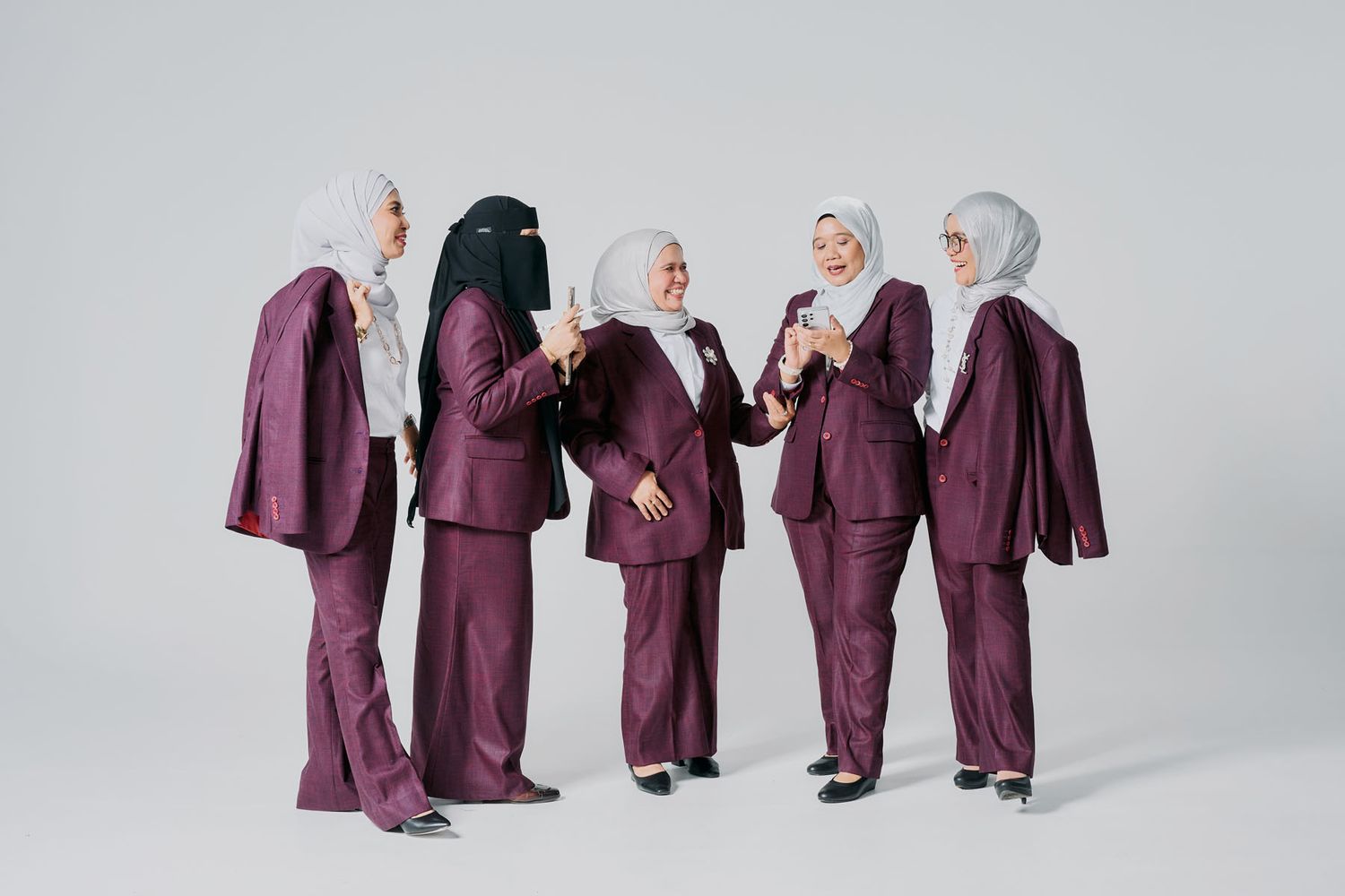 Five women in matching burgundy suits and hijabs pose together against a light gray background.