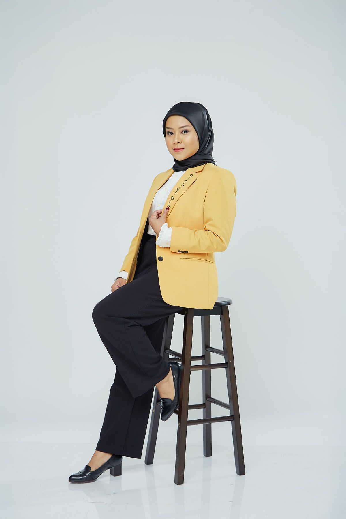 Young Muslim woman in yellow blazer and black hijab sitting on wooden stool against white background.