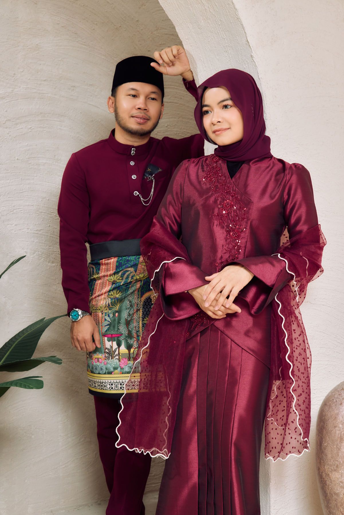 A Malaysian couple in coordinating deep red traditional outfits posing together under a white archway indoors.