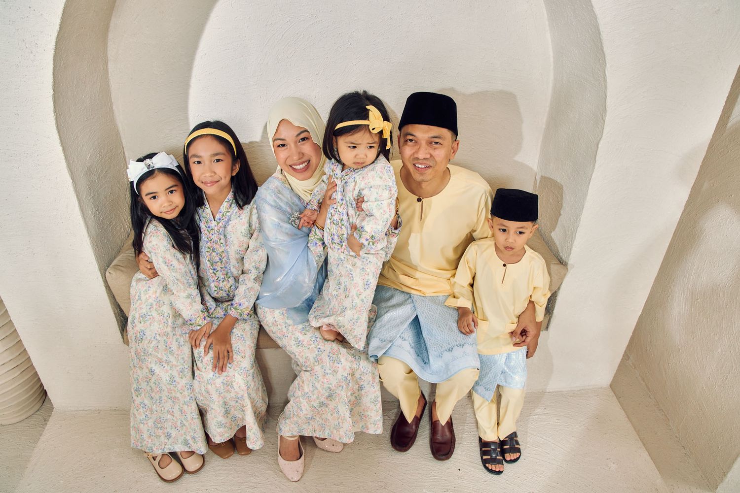 Overhead view of Malaysian Muslim family in pastel Hari Raya outfits seated in white arch.