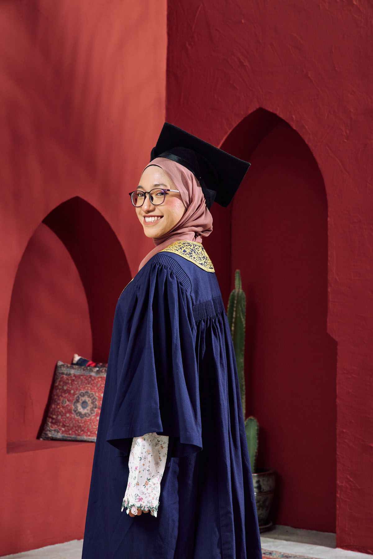 Smiling Muslim woman in navy graduation gown and mortarboard stands by red arched wall.