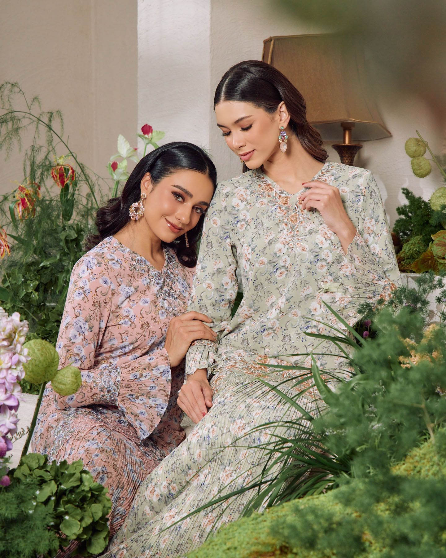 Two women in soft floral printed baju kurung dresses posing together among lush green tropical plants.