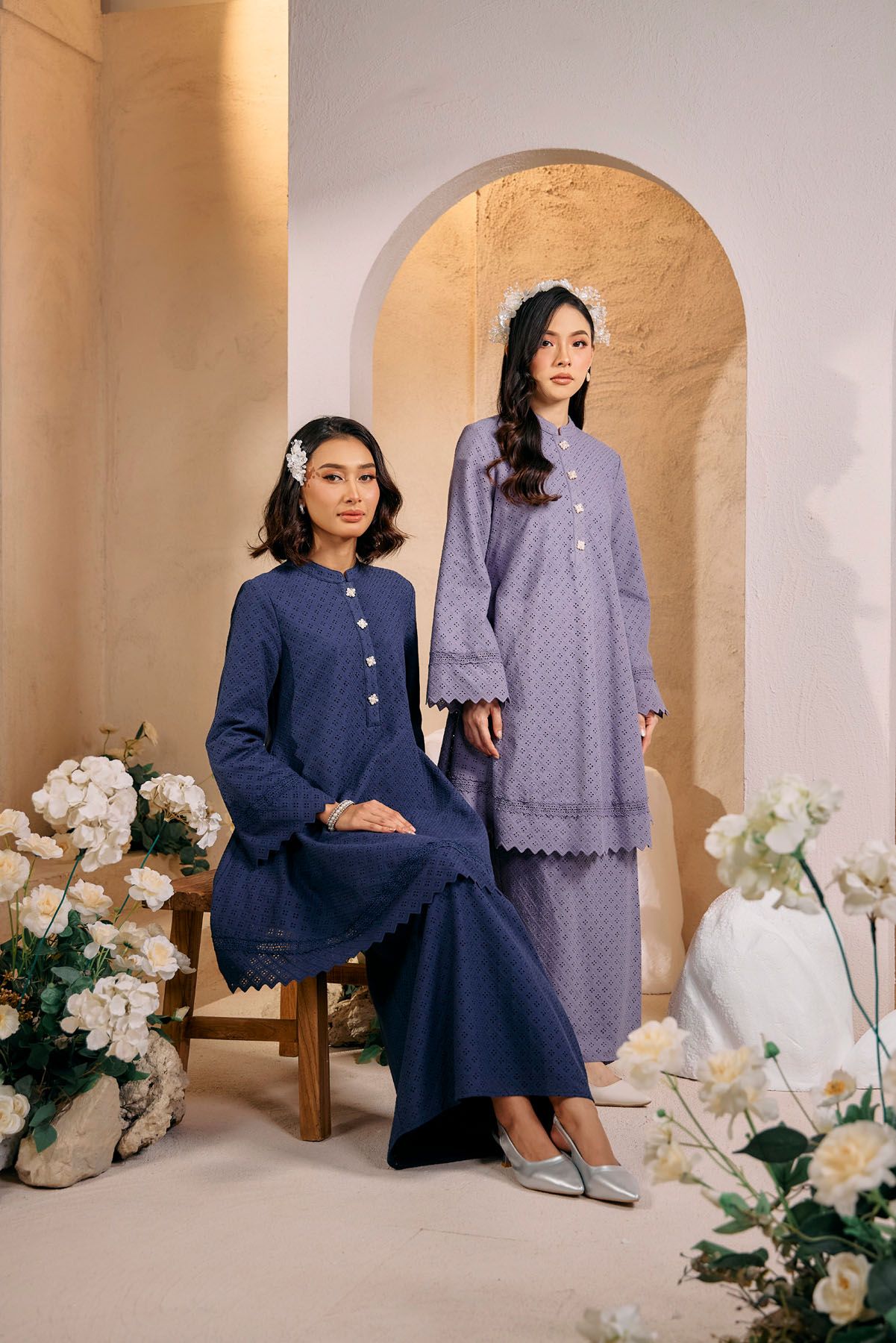 Two women modeling blue broderie anglaise baju kurung sets in an elegant arched interior setting.