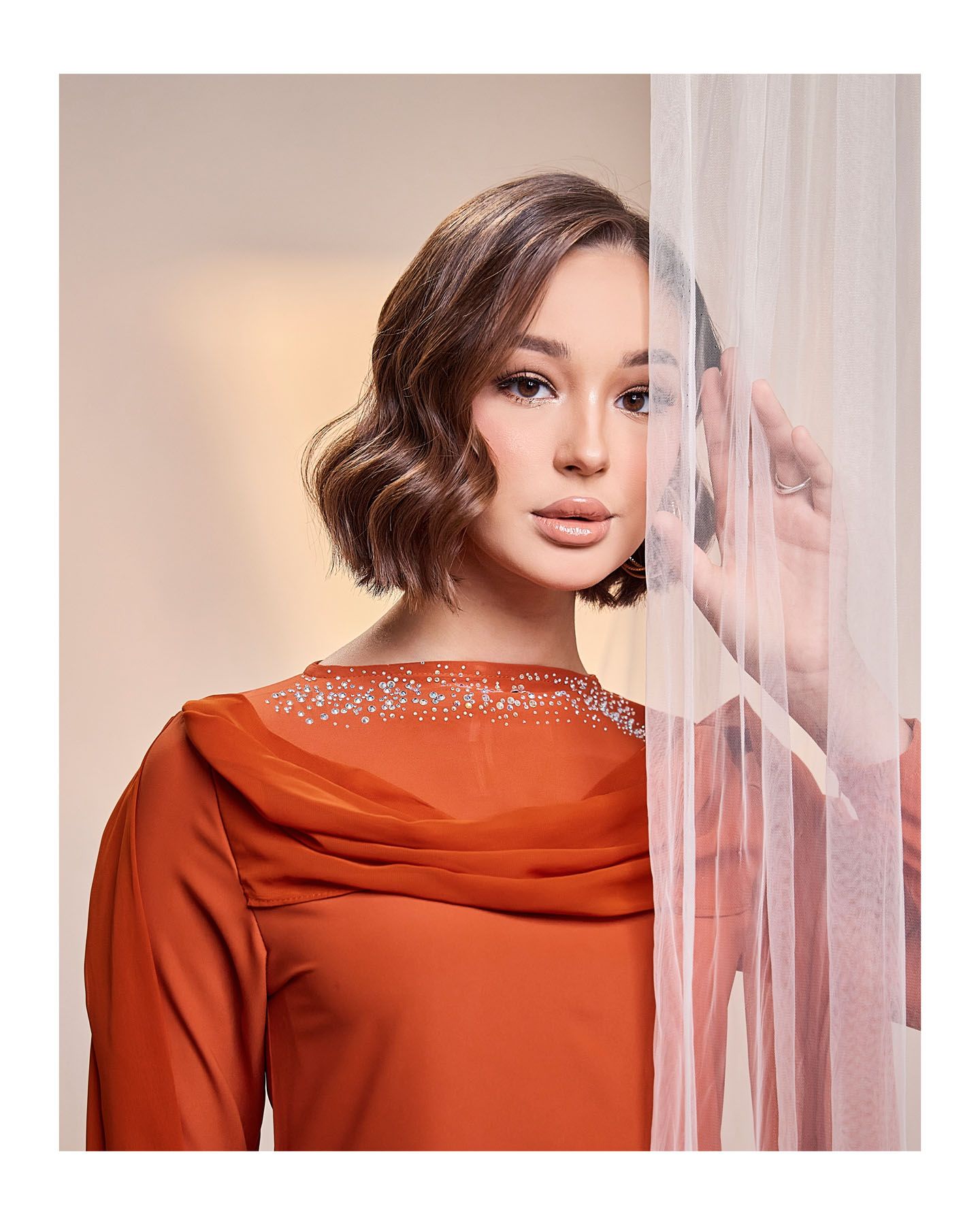 Woman in rust orange embellished neckline dress posing behind a sheer white curtain.