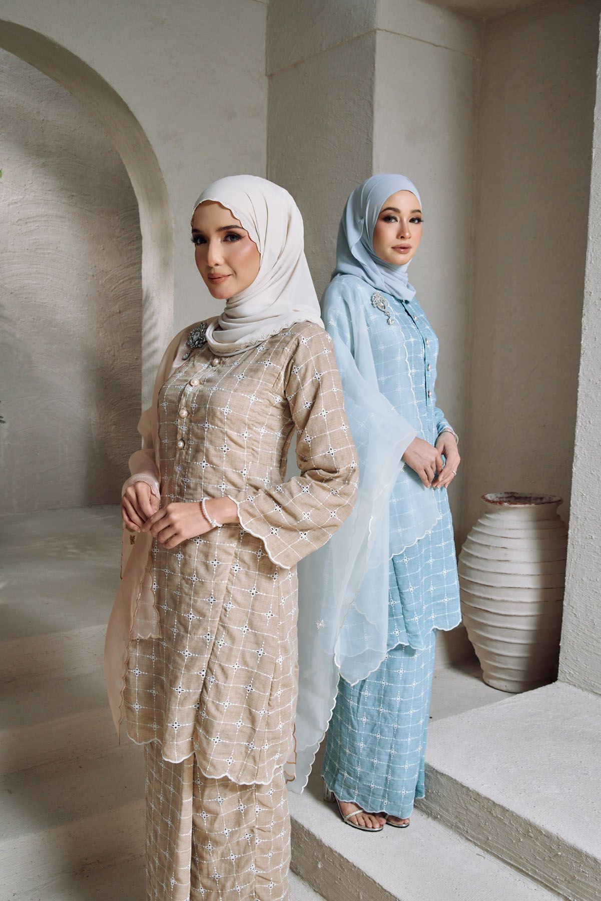 Two women in hijabs wearing elegant matching baju kurung sets in beige and light blue with scalloped details.