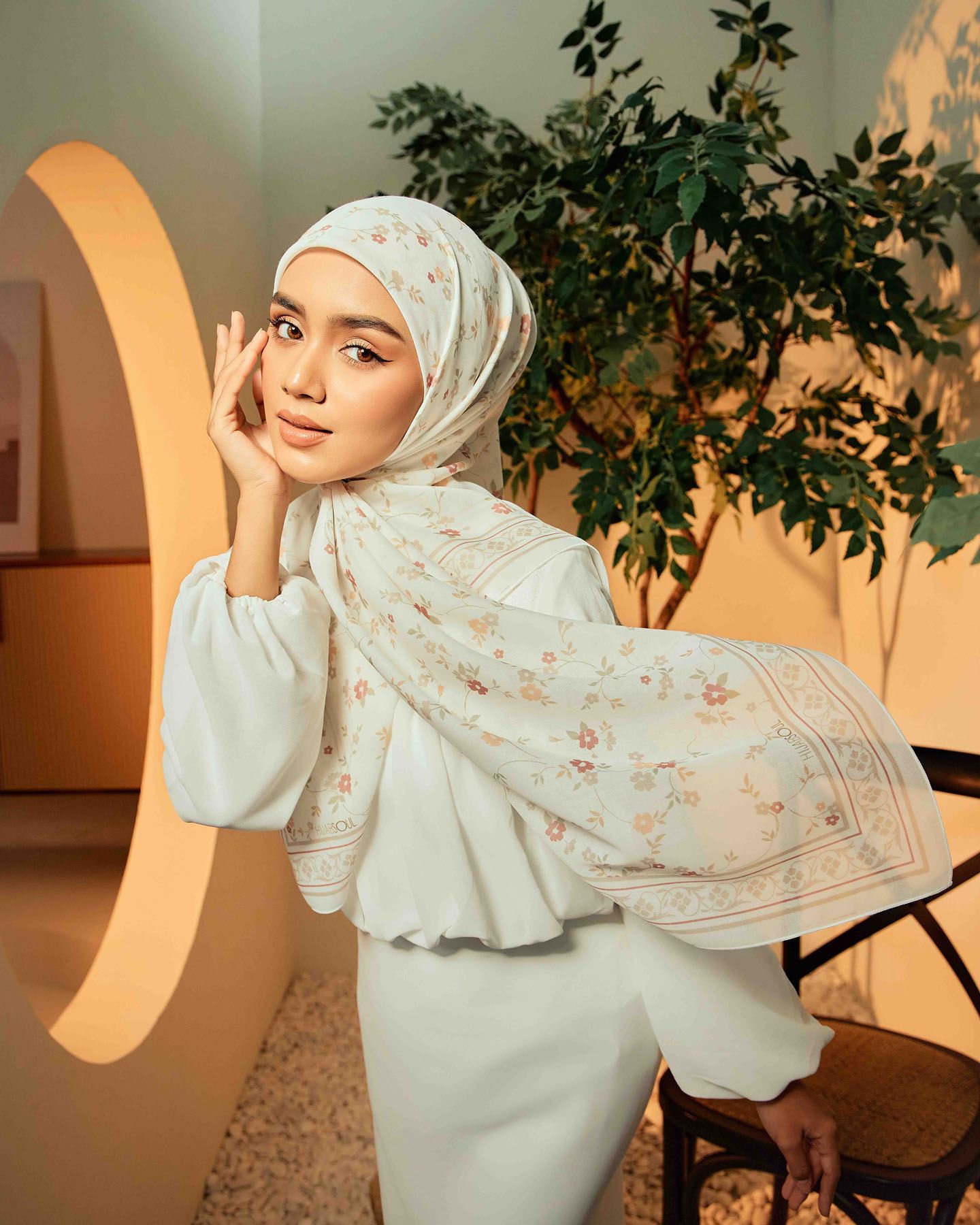 A woman in a floral white hijab and white outfit poses gracefully against a warm circular backdrop with greenery.