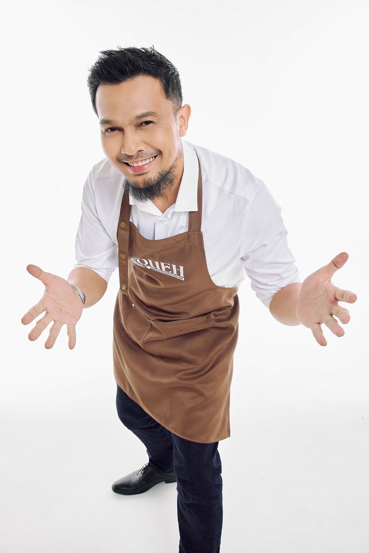 Smiling Asian man in brown apron leaning forward with open arms in welcoming gesture.