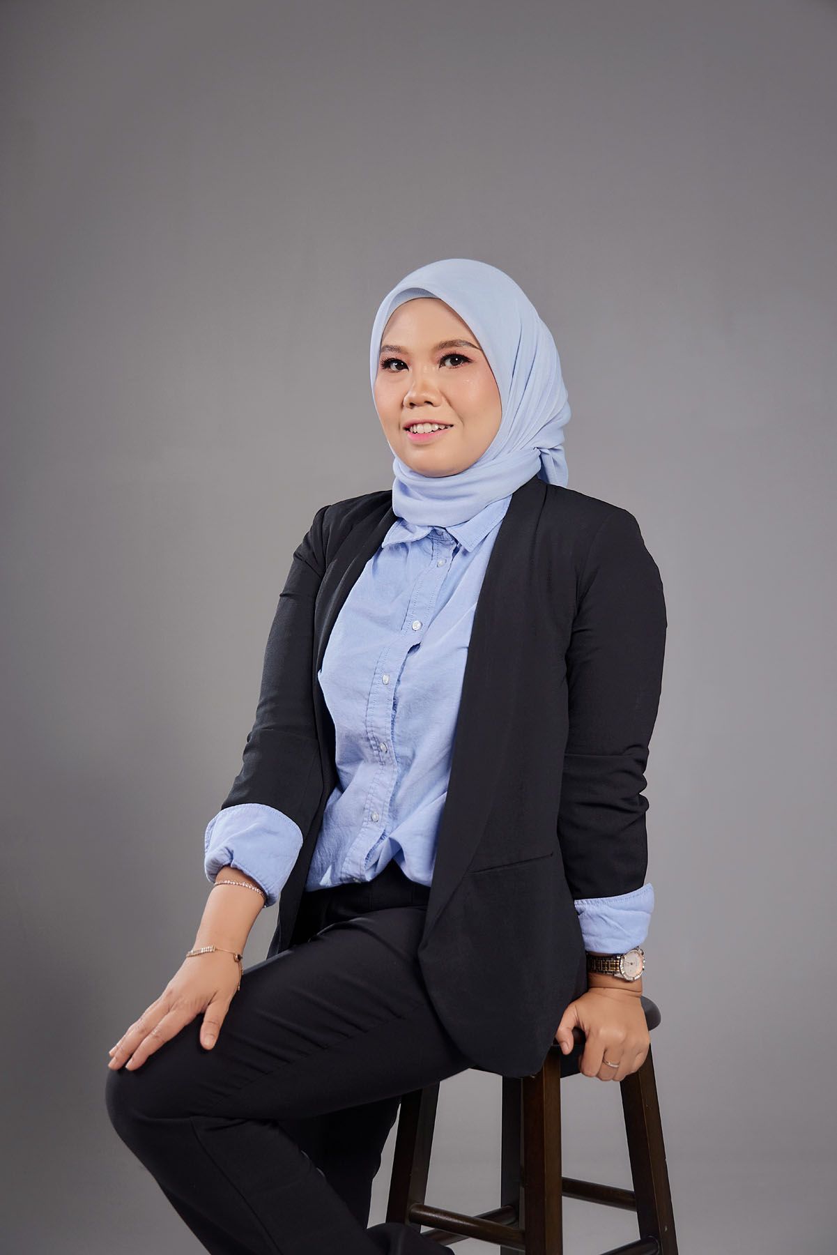 Professional Muslim woman in blue hijab and black blazer sitting on stool against gray background.