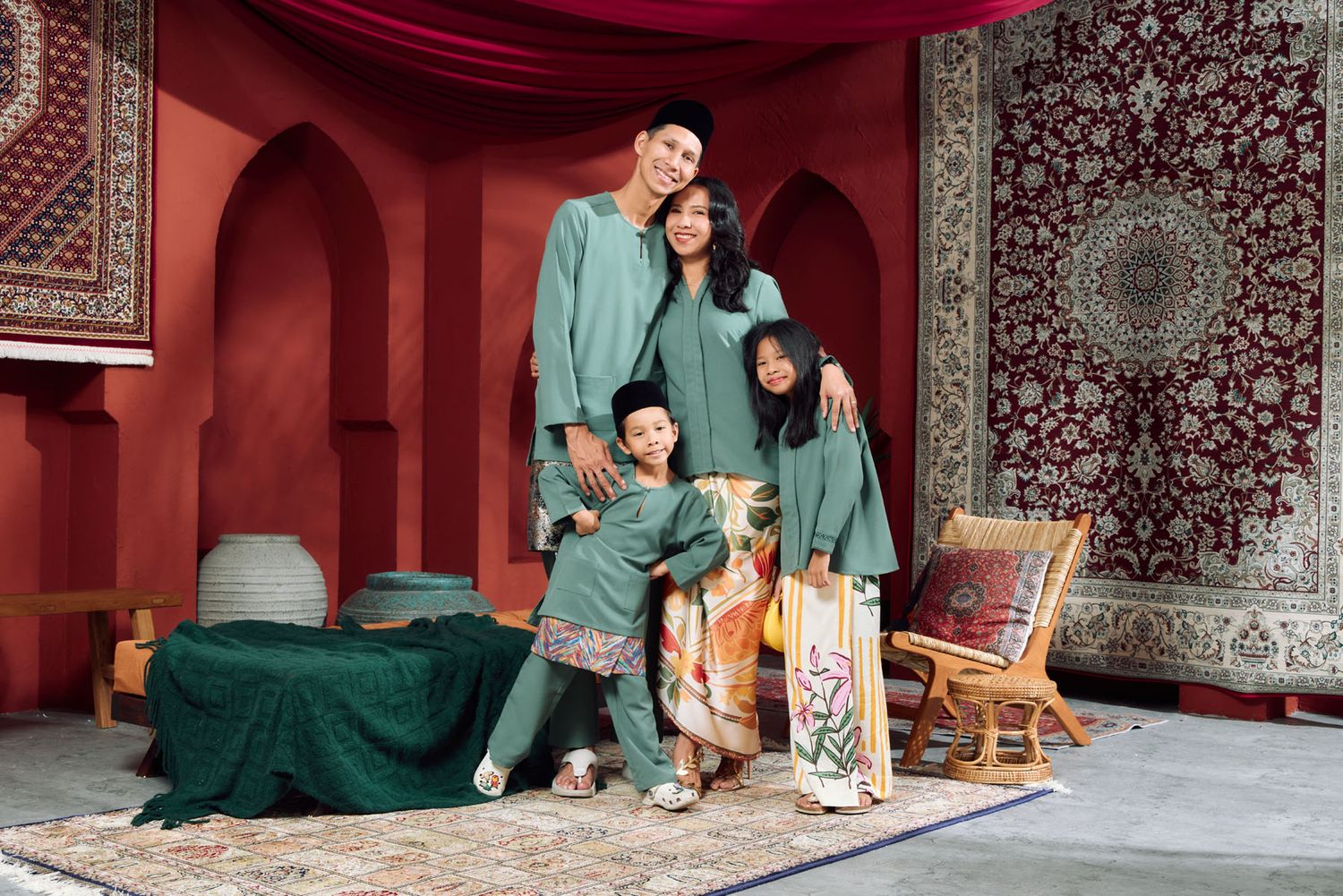 Family of three posing in coordinating dark green Baju Melayu and Baju Kurung in a red Moroccan-themed studio.