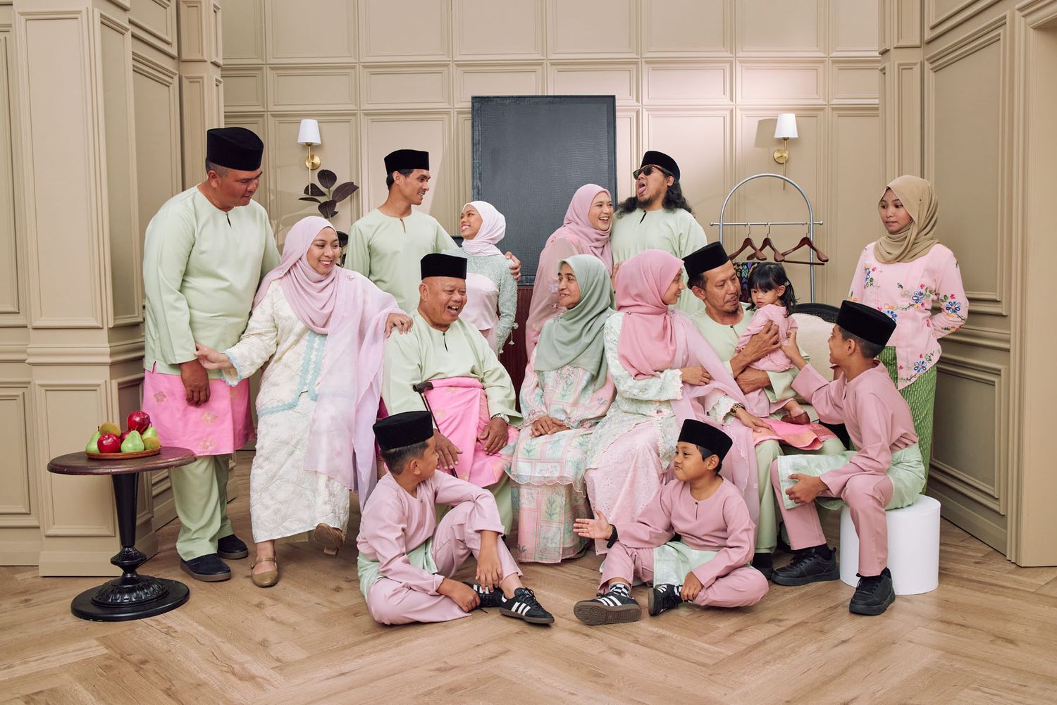 Large extended Malaysian family in pastel pink and green traditional Eid outfits in elegant indoor setting.