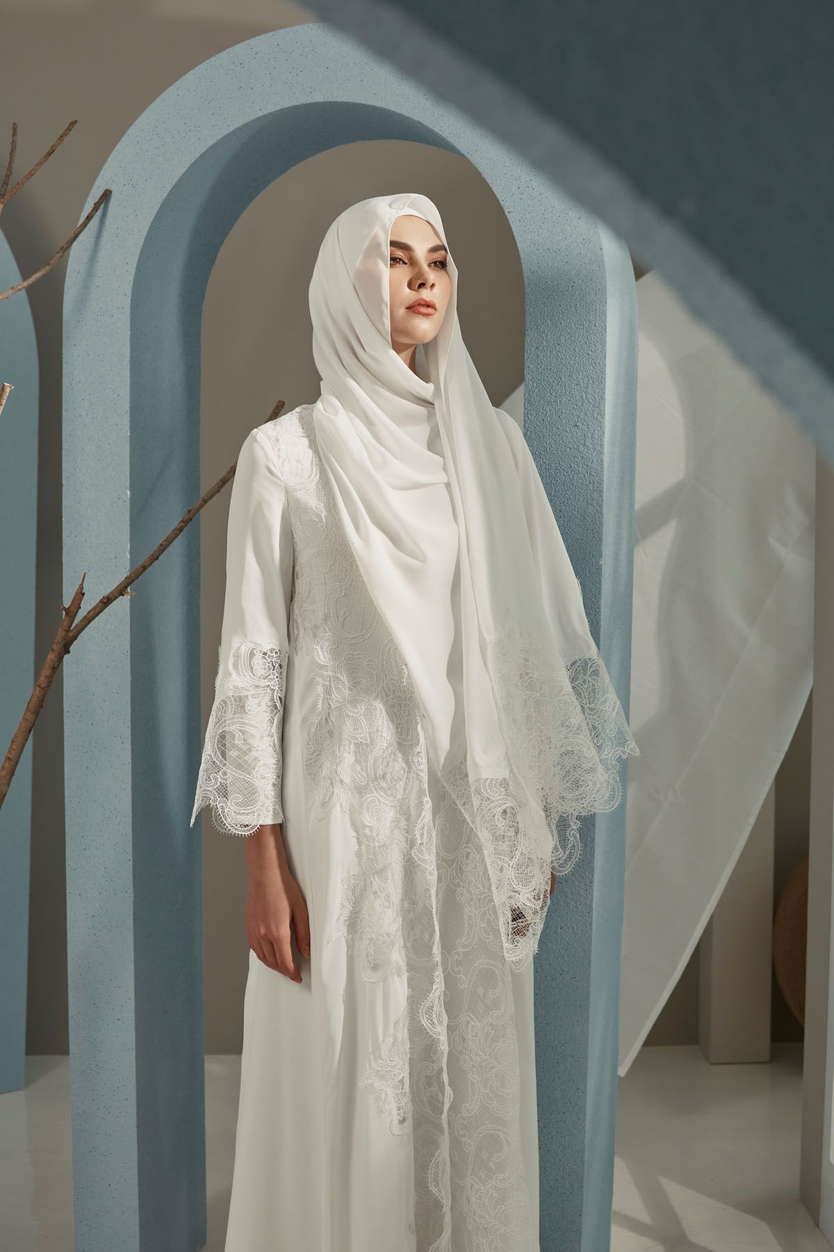 Woman in white hijab and lace-trimmed abaya stands serenely between blue arched doorways.