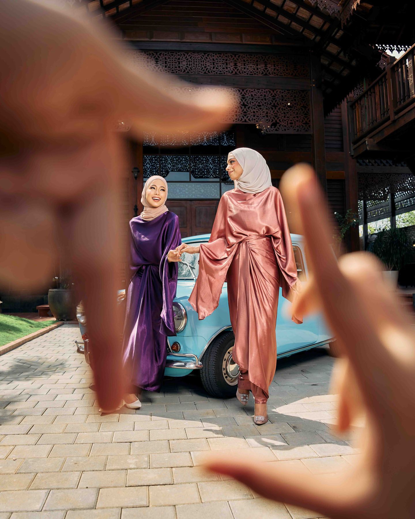 Two women in purple and pink silk hijab dresses pose near vintage blue car in wooden pavilion.