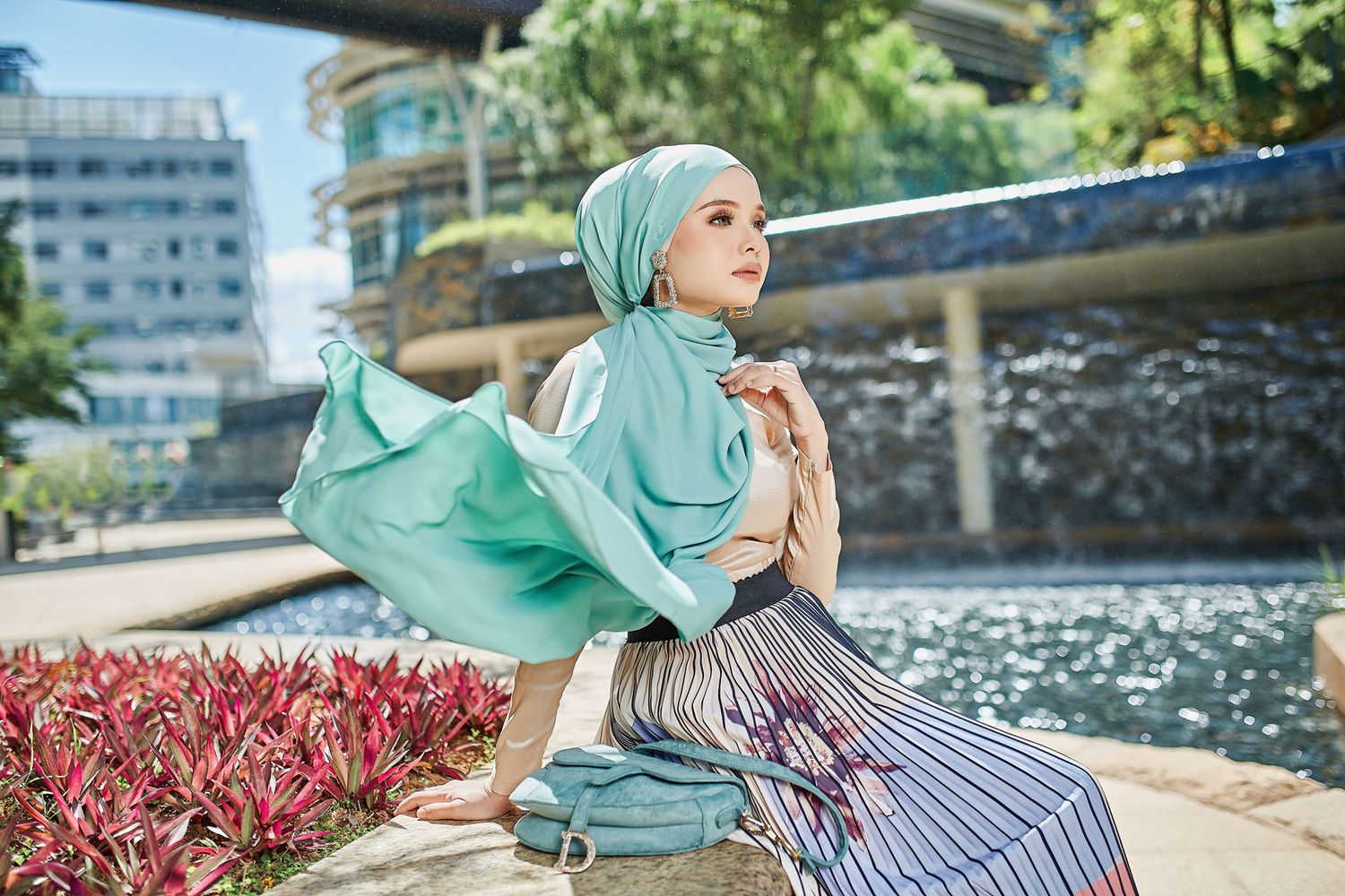 Woman wearing a mint green hijab with striped skirt seated outdoors near a urban waterway setting.
