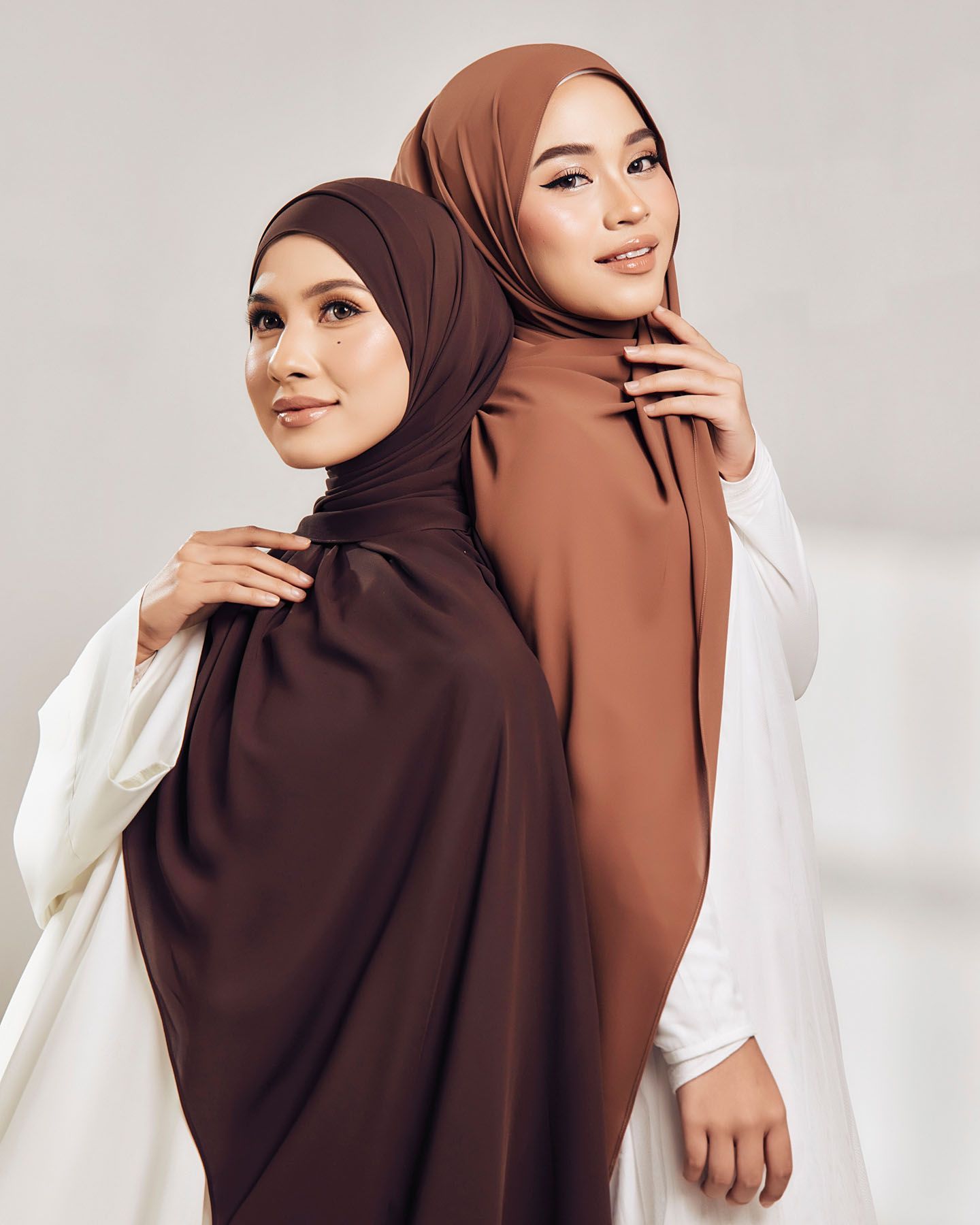 Two women wearing dark chocolate and caramel brown hijabs with white outfits in a minimalist studio setting.