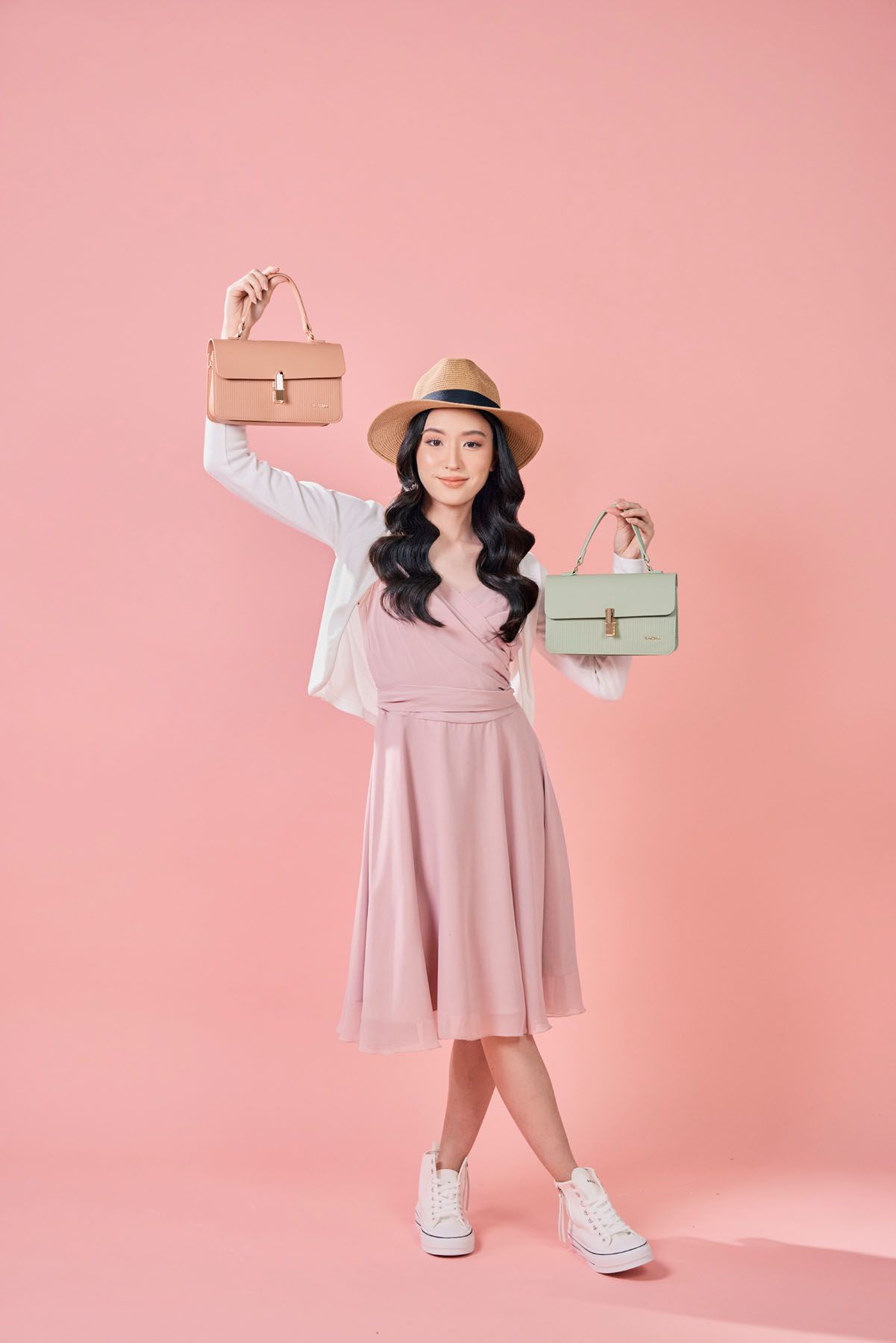 Asian woman in pink dress and hat holding up two small structured handbags in tan and green.