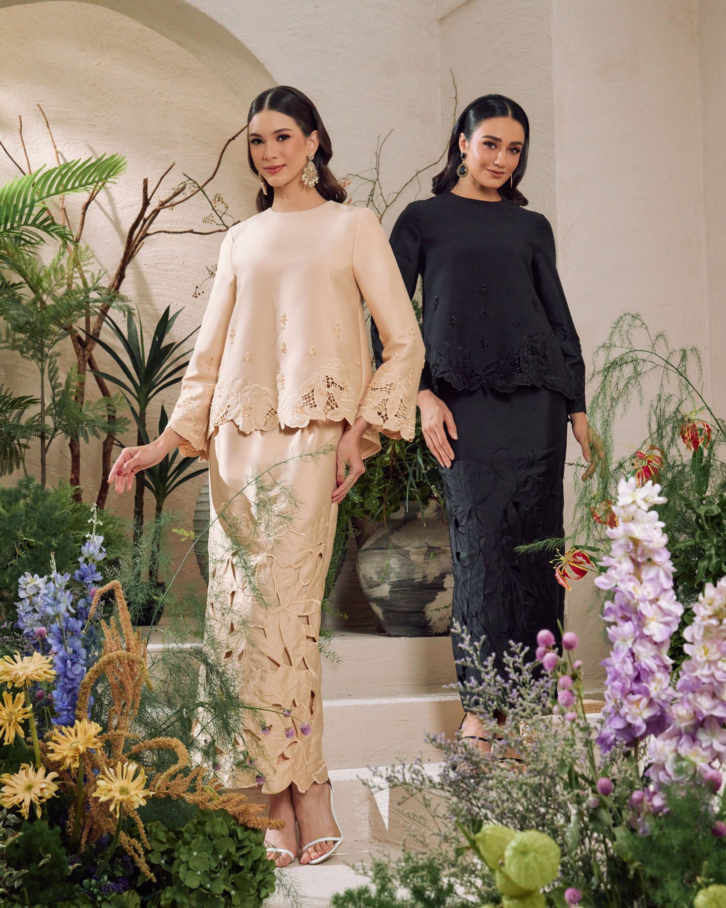 Two models wear elegant baju kurung, one in cream with floral cutwork and one in black lace, surrounded by flowers.