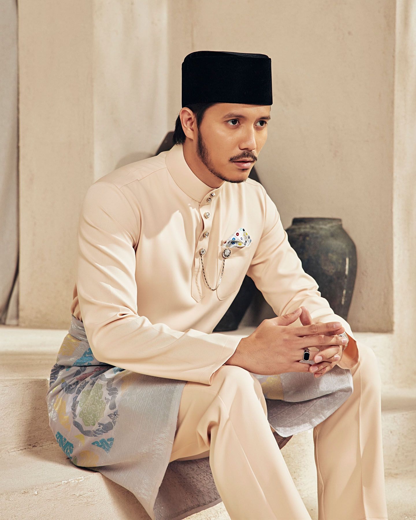 Man in cream baju melayu with black songkok and floral sampin sits gracefully.