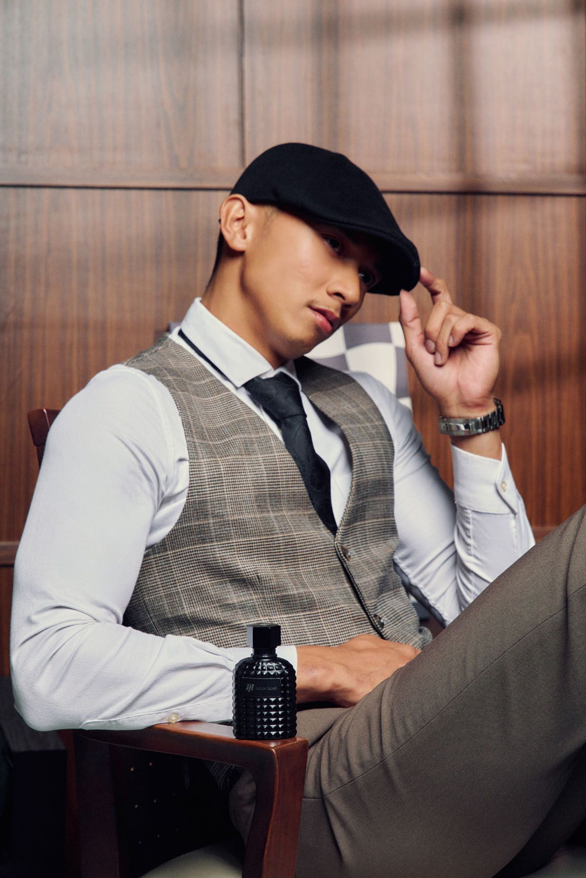 Stylish young man in plaid vest and flat cap seated with black fragrance bottle in wood-paneled room.
