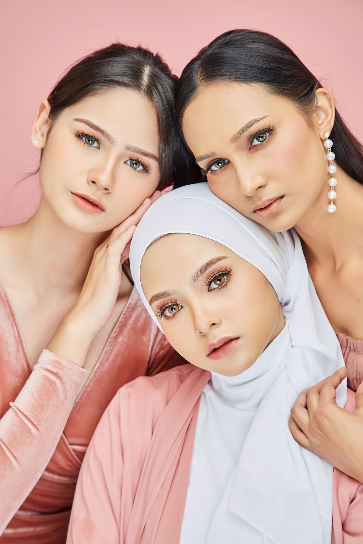 Three glamorous women with green eyes posing together against a pink background for a beauty shoot.